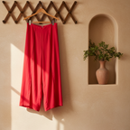Bright red wide-leg cropped pants hanging from a wooden hanger on a textured beige wall, with a decorative wooden lattice and a recessed shelf with a vase of greenery in the background.