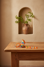 A colorful woven bird-shaped toy with a long tail rests on a wooden table in a warm, rustic room. In the background, a terracotta vase with green leaves sits in a recessed wall niche.