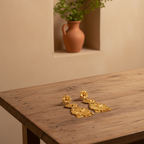 A pair of ornate gold-toned statement earrings displayed on a rustic wooden table, with a terracotta vase and green foliage in the background against a warm beige wall.
