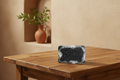 A rectangular slate soap bar with 'Hand Made' embossed on the front, resting on a wooden table in a warm, rustic interior setting with a terracotta vase in the background.