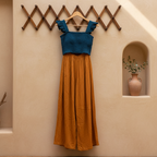 A two-piece dress with a textured teal blue crop top and a flowing mustard yellow skirt, hanging on a wooden hanger from a decorative wall rack against a beige stucco wall.