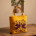 A yellow canvas tote bag with a dark purple graphic of two birds and leaves, displayed on a wooden table with a beige wall and a potted plant in the background.