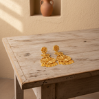 A pair of ornate gold jhumka earrings with tassel fringe, displayed on a rustic wooden table in front of a beige stucco wall with a built-in niche holding a small terracotta vase with green stems.