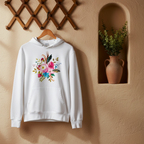 A white pullover hoodie hanging on a wooden hanger against a warm beige wall, featuring a colorful floral and feather graphic on the chest. The setting includes a wooden lattice wall decor piece and a recessed niche with a terracotta vase containing green plants.